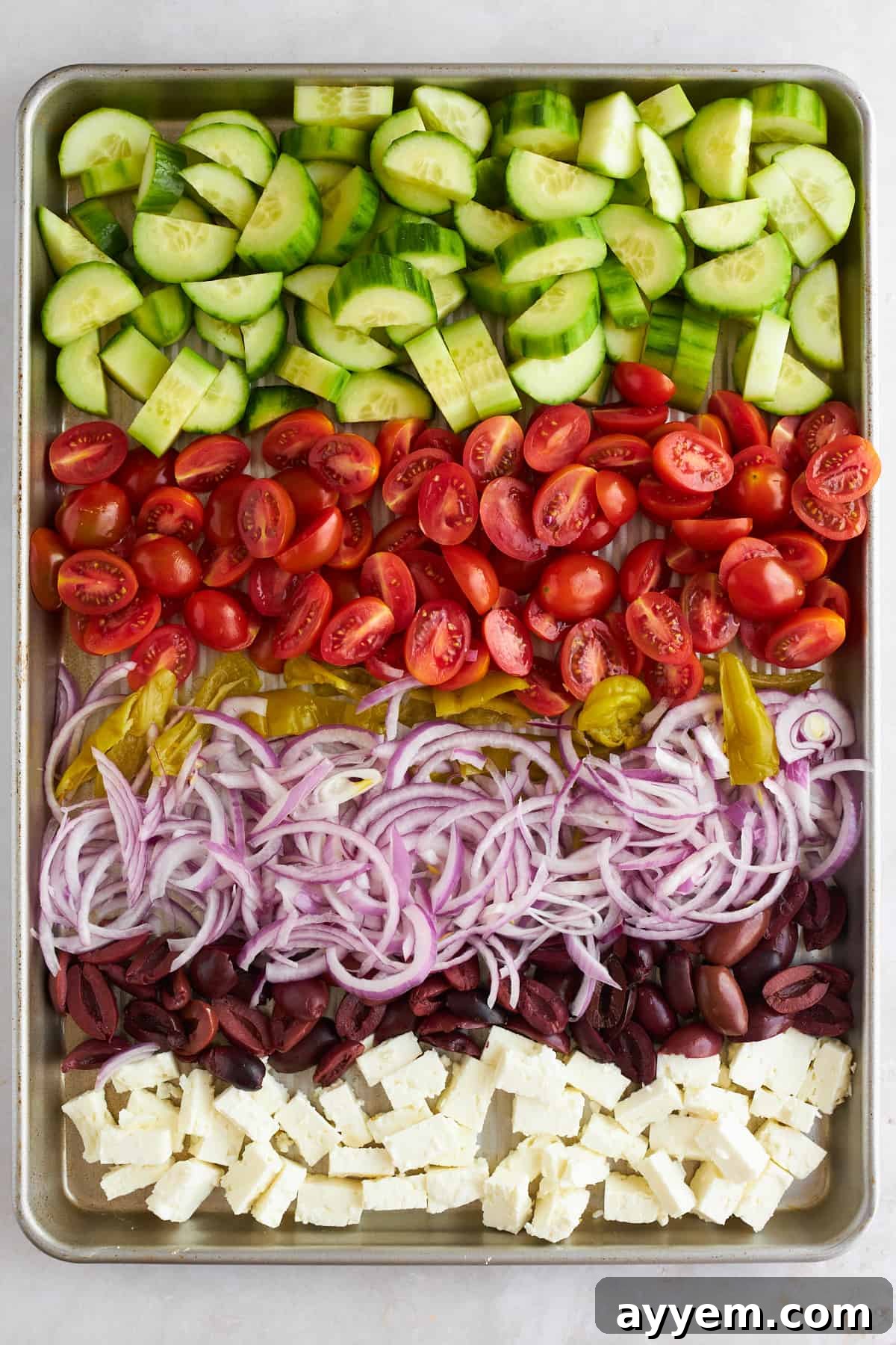 Cucumbers, tomatoes, pepperoncini peppers, red onions, and olives on a baking sheet. 