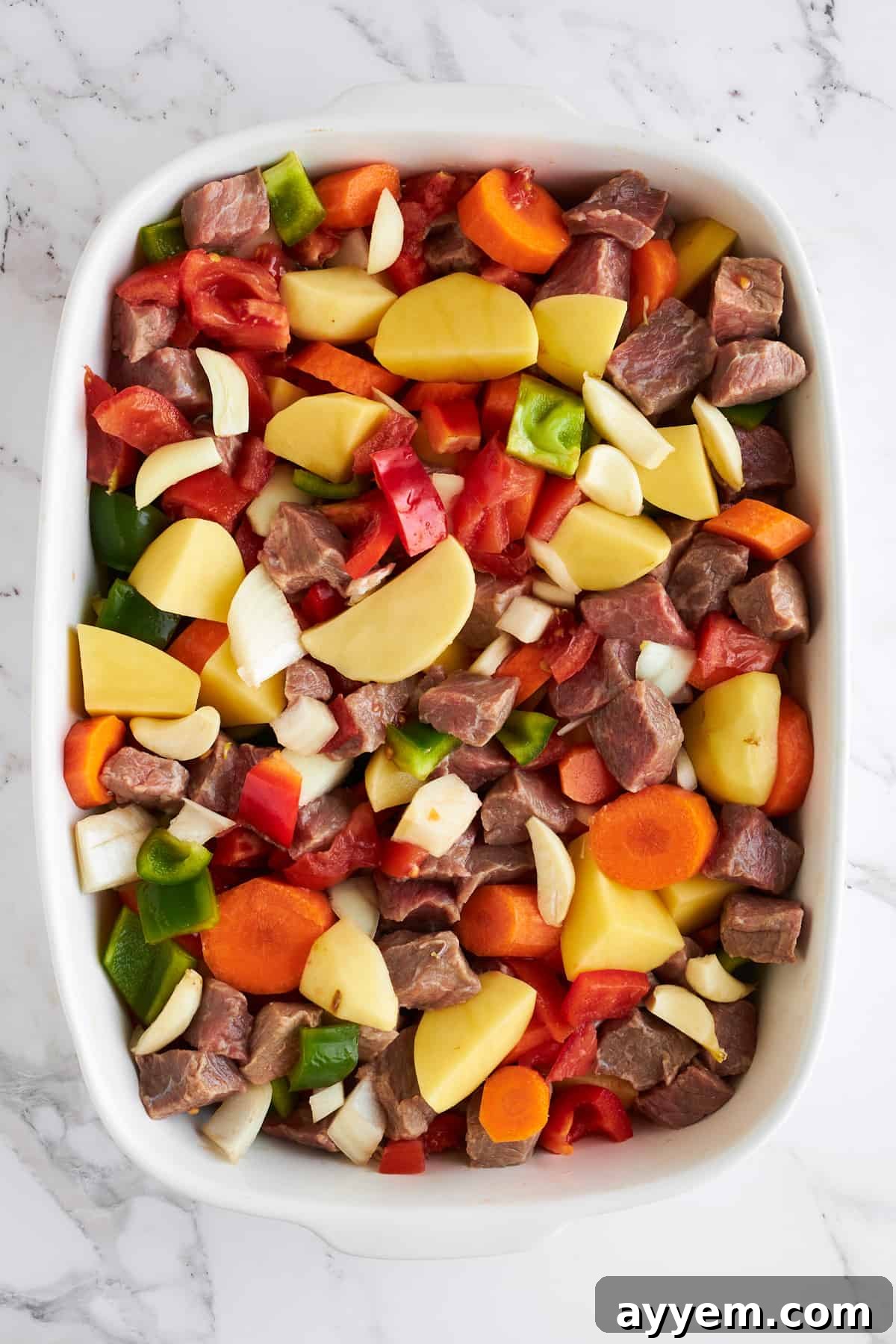 Uncooked beef cubes and a vibrant mix of fresh vegetables artfully arranged in a large baking dish, ready for seasoning and sauce.