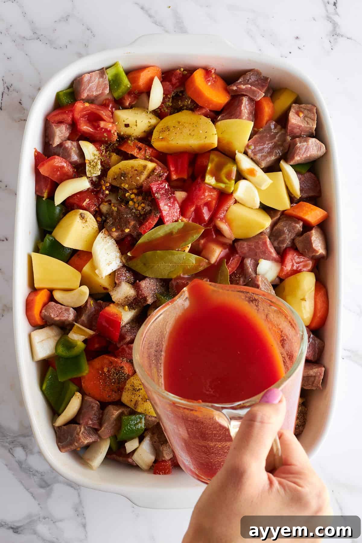 A rich, reddish-brown sauce being steadily poured over the prepared beef and vegetables in a baking dish, ready to be covered and baked.