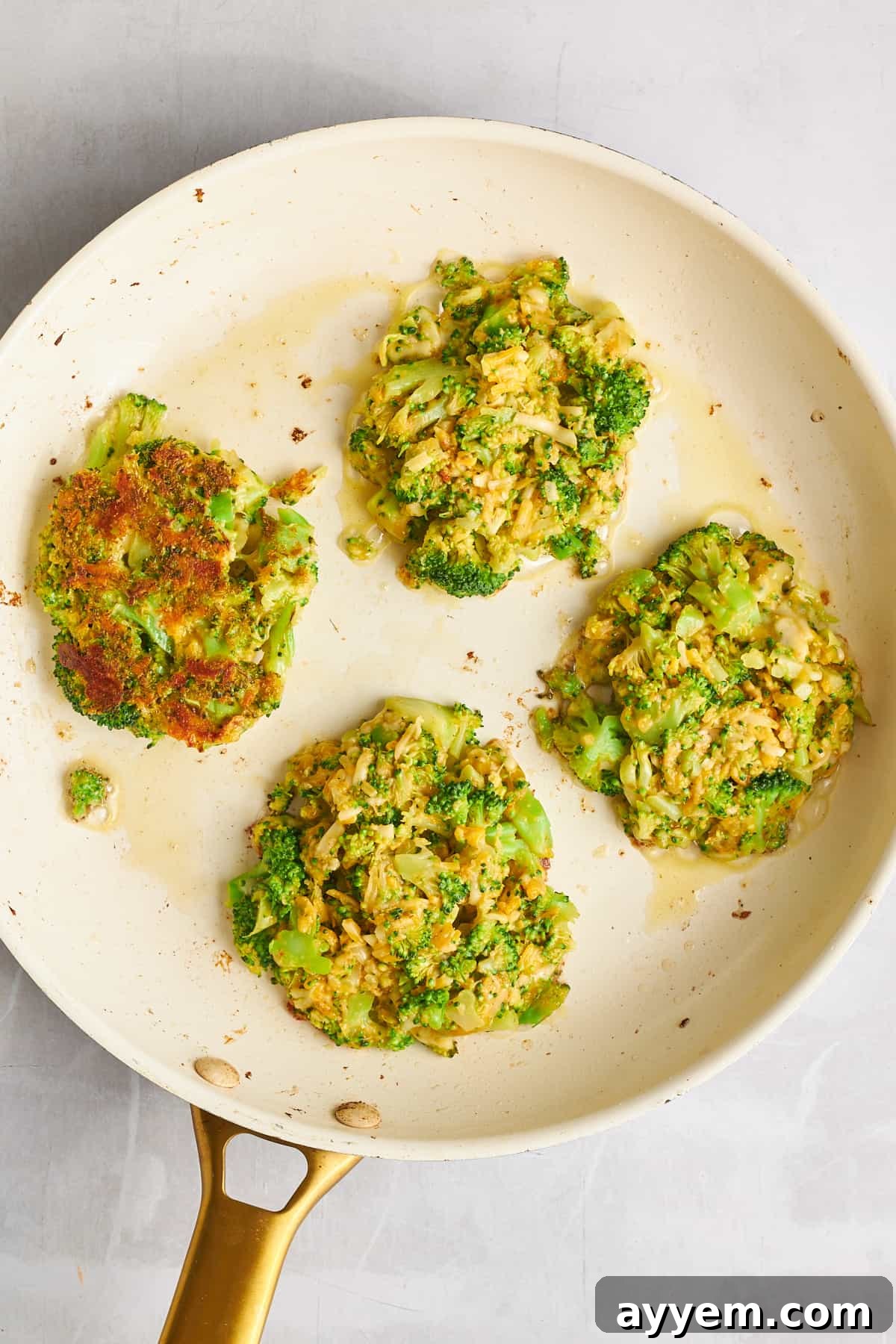 Four cheesy broccoli fritters sizzling in a non-stick skillet, turning golden brown.