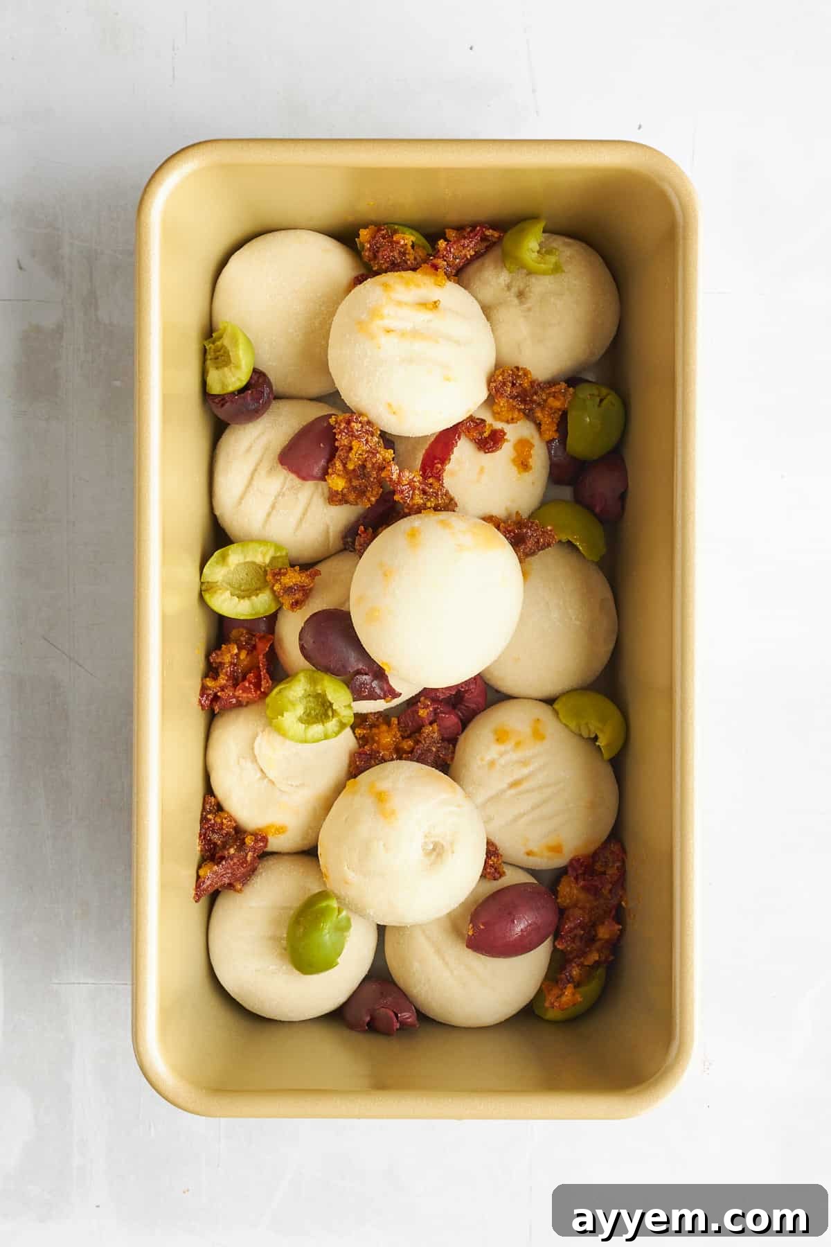 Raw bread rolls, olives, and sun-dried tomatoes arranged in a loaf pan, ready for rising and baking.