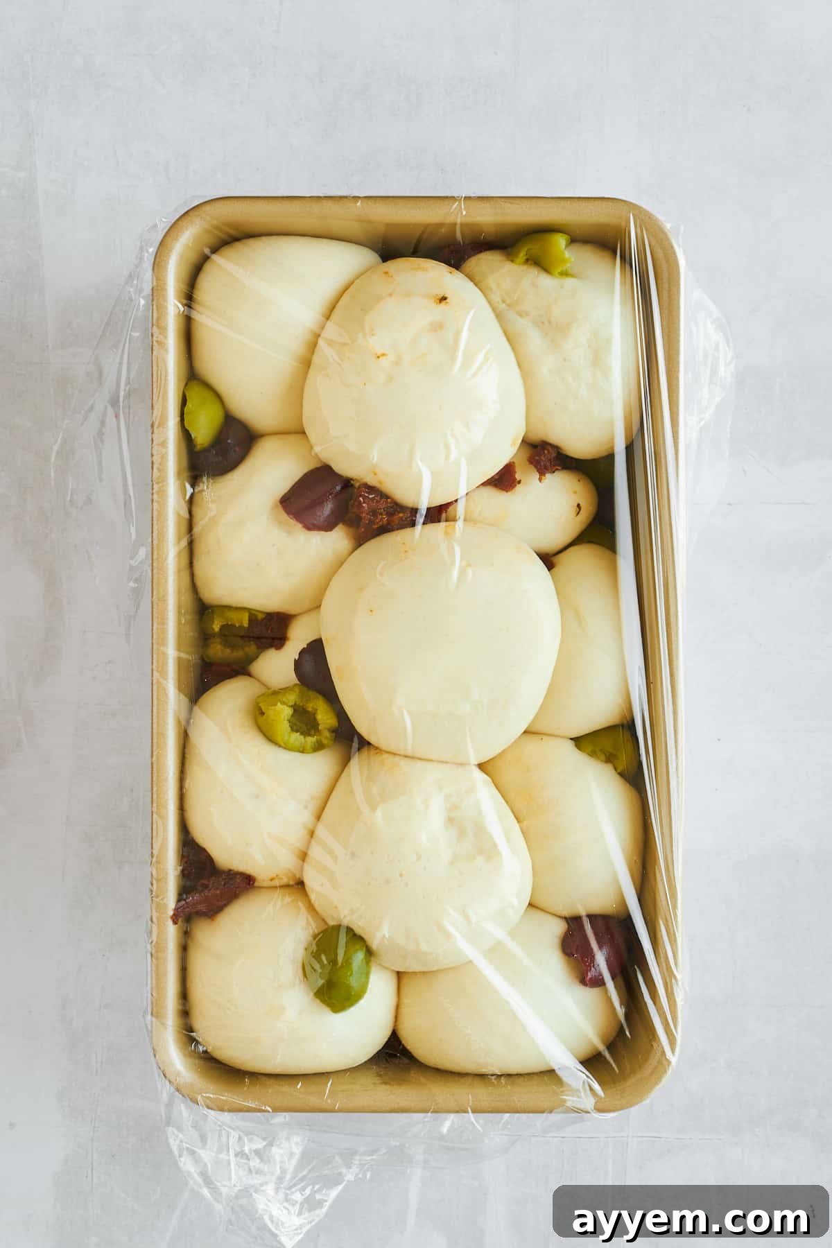 Raw bread rolls, olives, and sun-dried tomatoes in a loaf pan, covered with plastic wrap, showing the rising process.