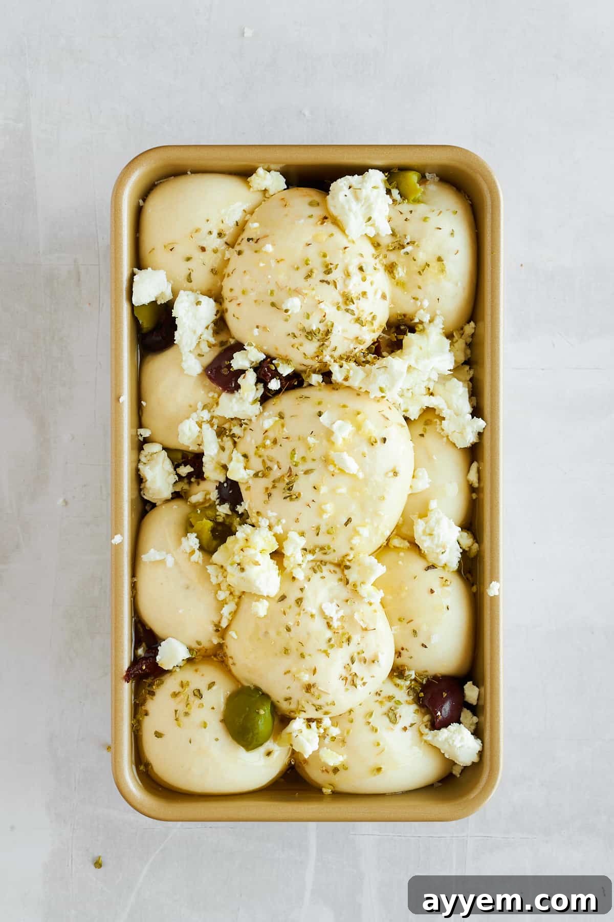 Risen bread rolls, layered with olives, sun-dried tomatoes, and feta cheese, ready for the oven in a bread pan.