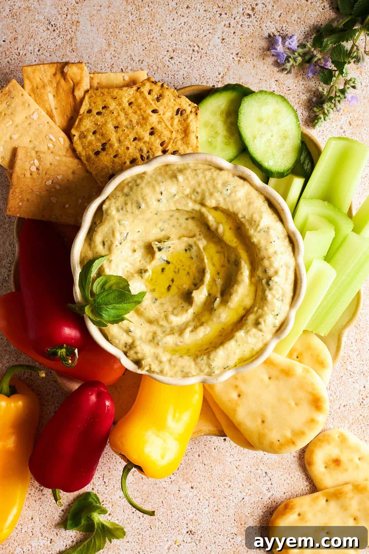Creamy Herb Zucchini Dip 3 A vibrant and fresh zucchini dip in a rustic bowl, nestled on a wooden platter alongside an array of colorful dippers including crunchy cucumber spears, crisp celery sticks, golden pita triangles, miniature sweet bell peppers in red and yellow, and various artisanal crackers, illustrating a perfect healthy snack or appetizer spread.