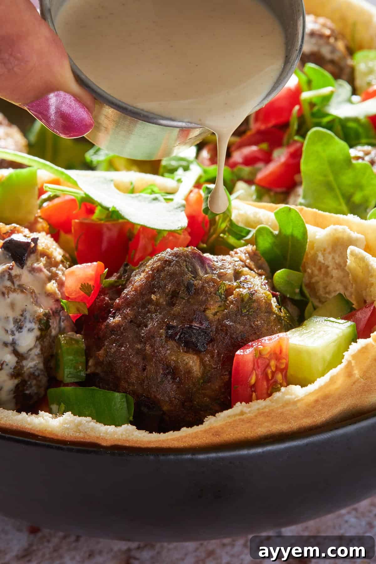 Creamy tahini sauce generously drizzled over a warm pita pocket, overflowing with juicy Mediterranean beef meatballs and a fresh, colorful salad. 