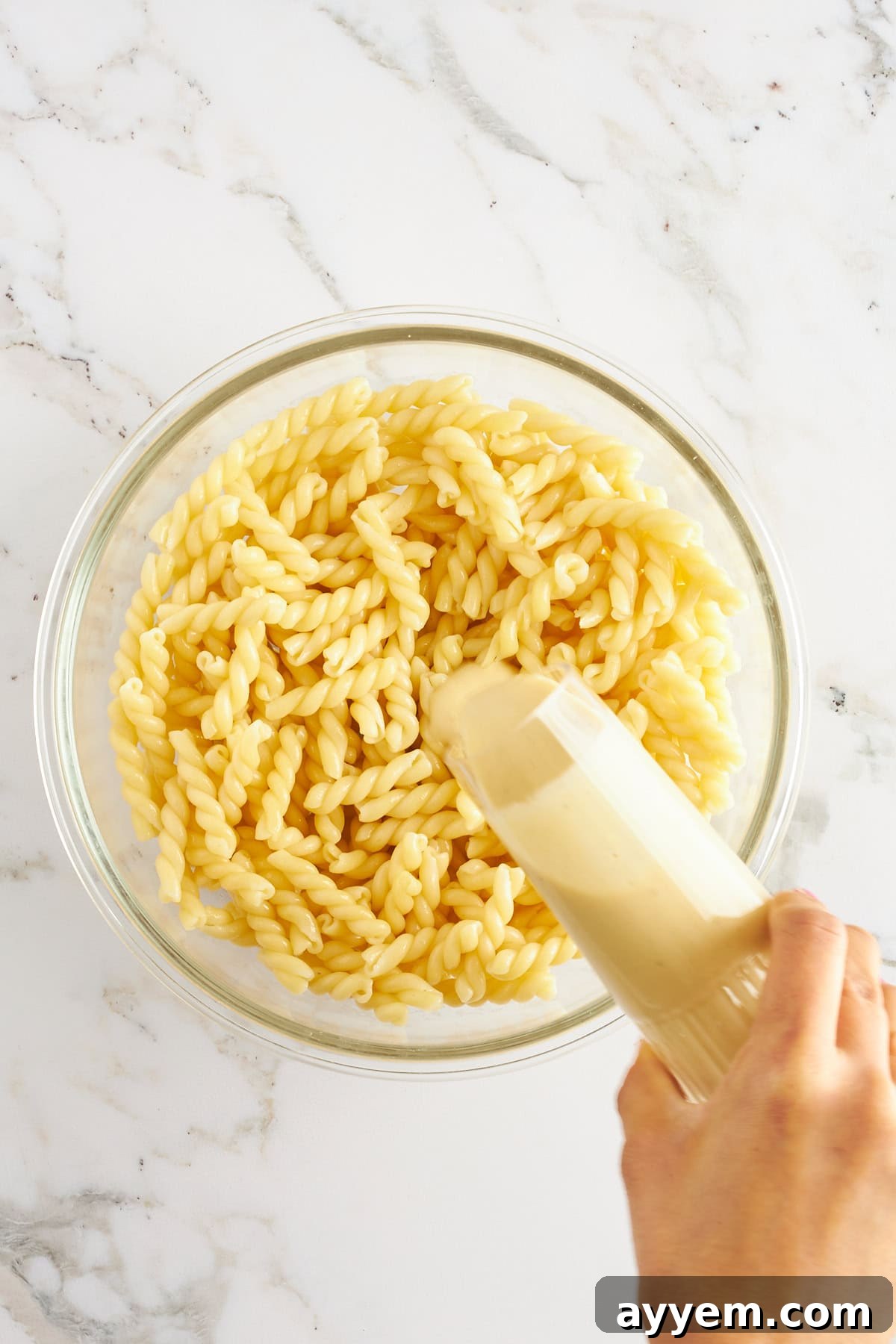 Zesty Caesar Pasta Bowl 3 A bowl of medium-sized pasta noodles and a hand pouring Caesar dressing on top.