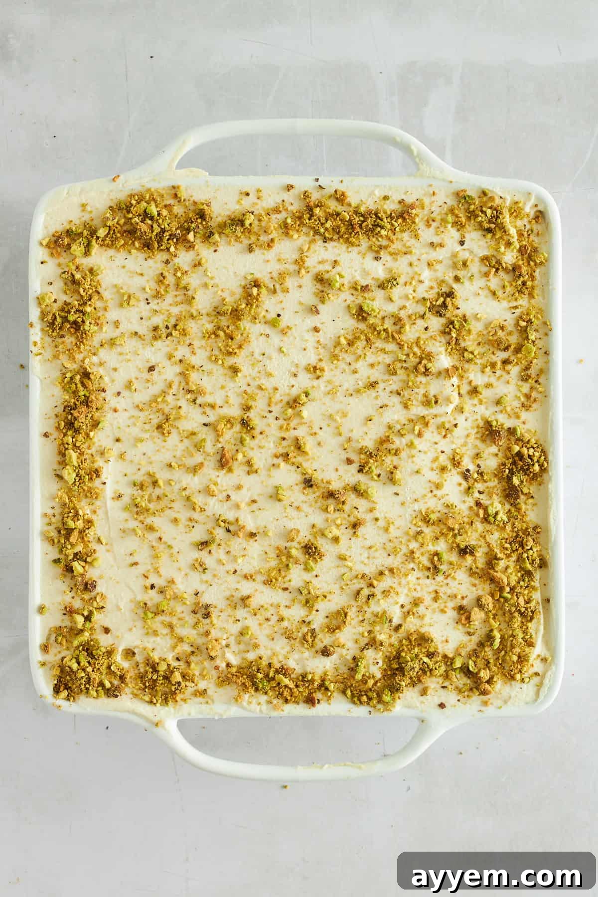 A pristine square baking dish filled with homemade pistachio tiramisu, ready to be chilled and served, showing the smooth, light green top layer before garnish.