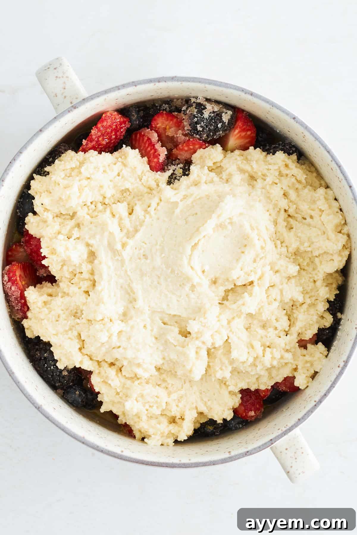 Uncooked mixed berry cobbler in a round baking dish, topped with a raw, fluffy biscuit dough and sprinkled with turbinado sugar, before entering the oven.