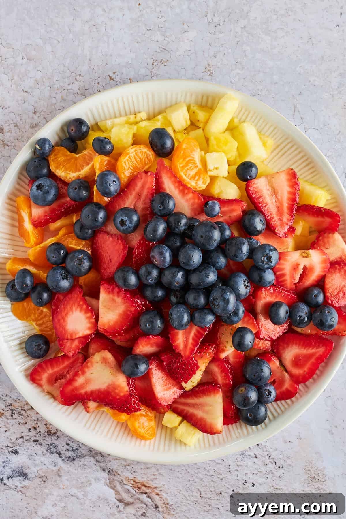 A vibrant bowl filled with an assortment of fresh fruits: ripe strawberries, plump blueberries, golden pineapple chunks, and bright mandarin orange segments, ready to be transformed into Ambrosia fruit salad.