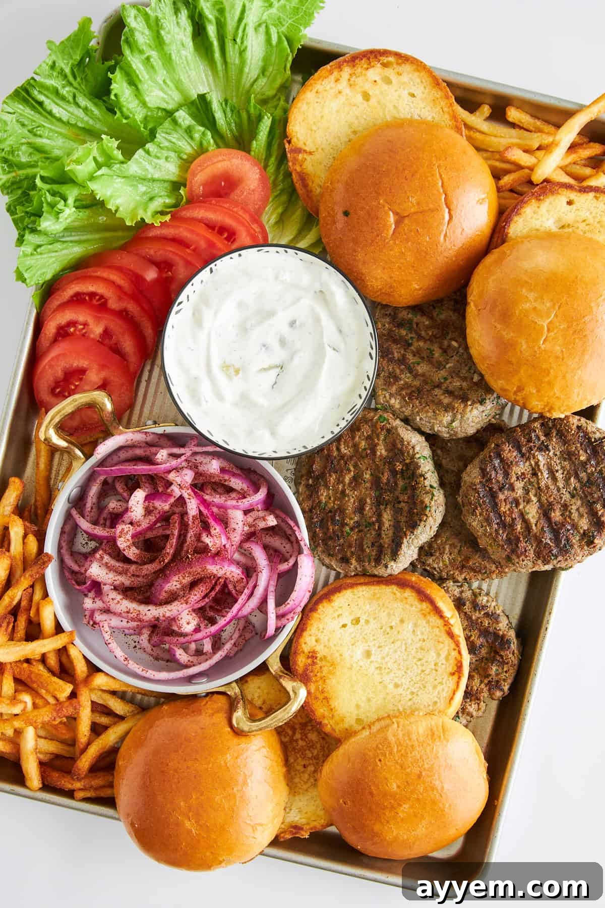 A vibrant platter of lamb kofta burger patties, toasted buns, fresh sumac onions, ripe tomatoes, crisp lettuce, and golden fries.