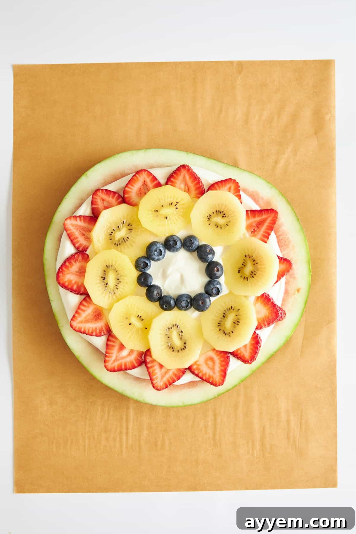 A watermelon pizza base beautifully topped with Greek yogurt, freshly sliced strawberries, bright green kiwi, and plump blueberries, creating a colorful pattern.