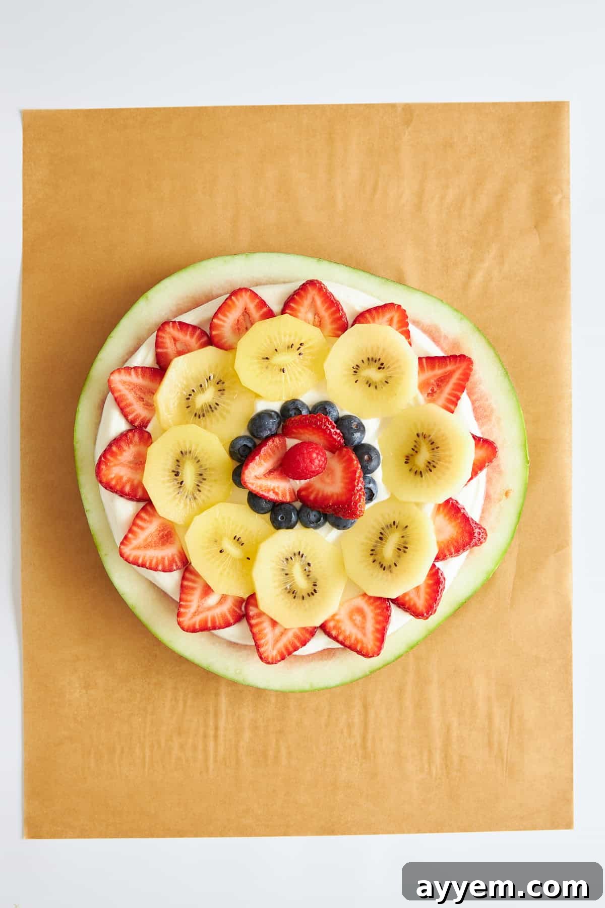 A vibrant watermelon pizza, fully assembled with a creamy yogurt base, mixed berries, and kiwi slices, ready for a final sprinkle of granola.