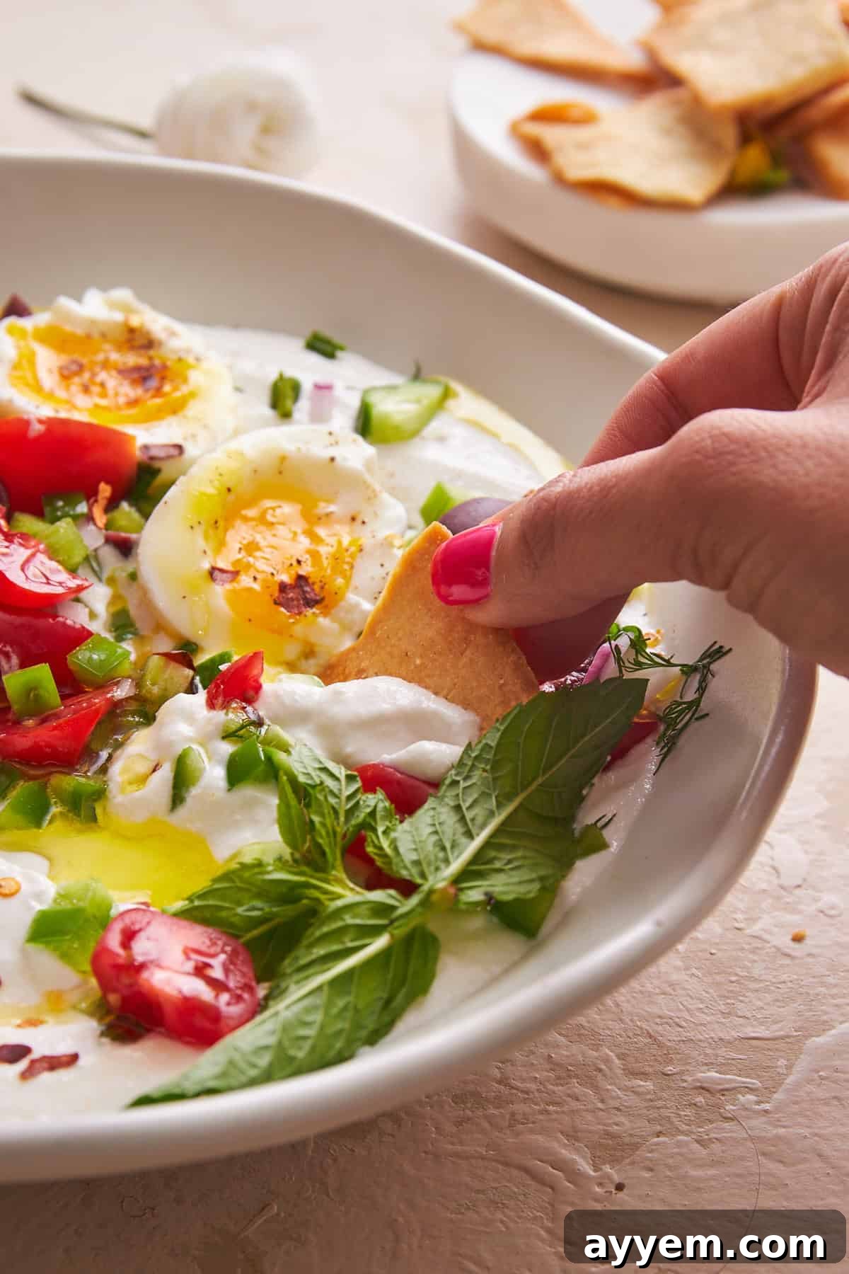 A hand gently dipping a crispy pita chip into a vibrant bowl of Mediterranean whipped cottage cheese salad, inviting a taste.