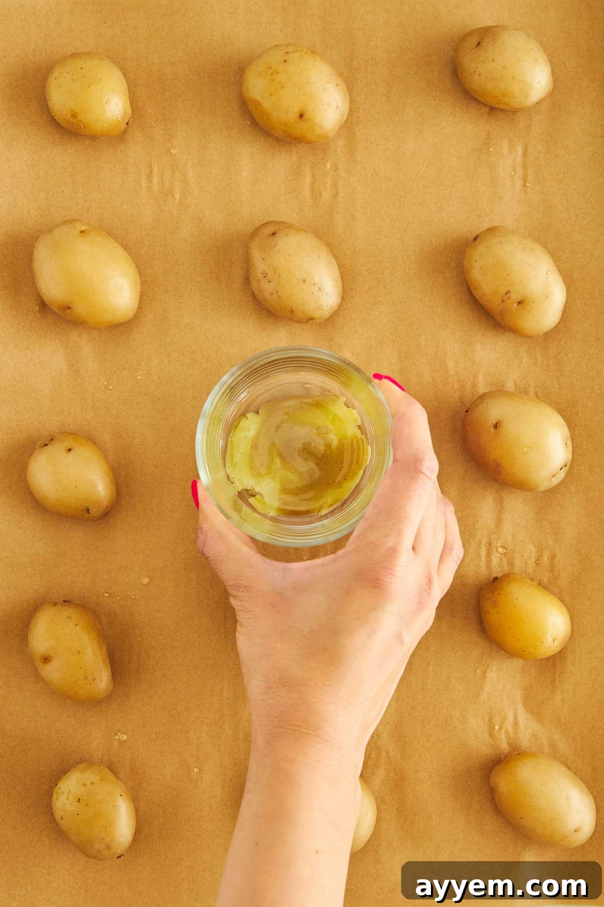 A hand pressing a glass cup down to smash a boiled potato on a baking sheet.