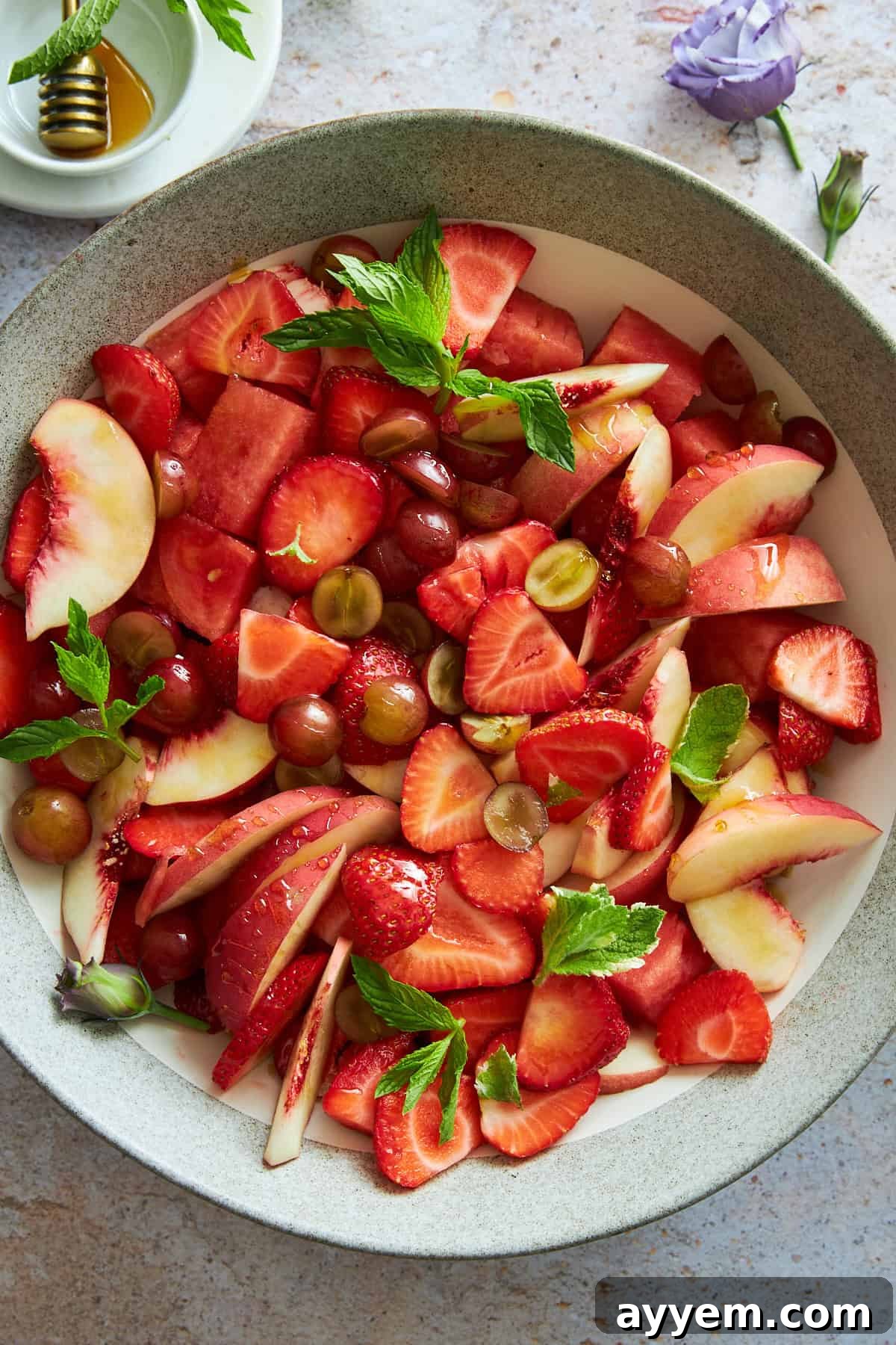 A close-up of a bowl of peach watermelon salad, featuring visible grapes, strawberries, and fresh mint leaves.