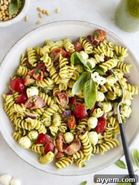A generous bowl of pasta salad with vibrant green pesto, sun-dried tomatoes, and fresh herbs, with a fork resting beside it.