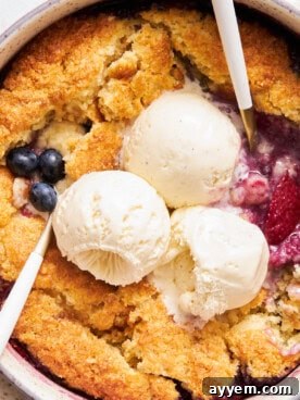 Close up view of mixed berry cobbler topped with three scoops of vanilla ice cream, showcasing its rich fruit filling and golden biscuit topping, ready to be enjoyed.