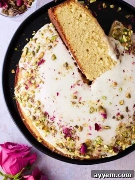 An elegantly decorated Persian Love Cake, infused with rose water and cardamom, topped with glaze, rose petals, and pistachios, with a slice removed.