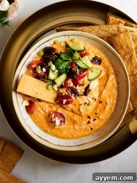 A rustic bowl of spicy feta dip, Tirokafteri, generously topped with chopped fresh vegetables and served with a crisp cracker for dipping.