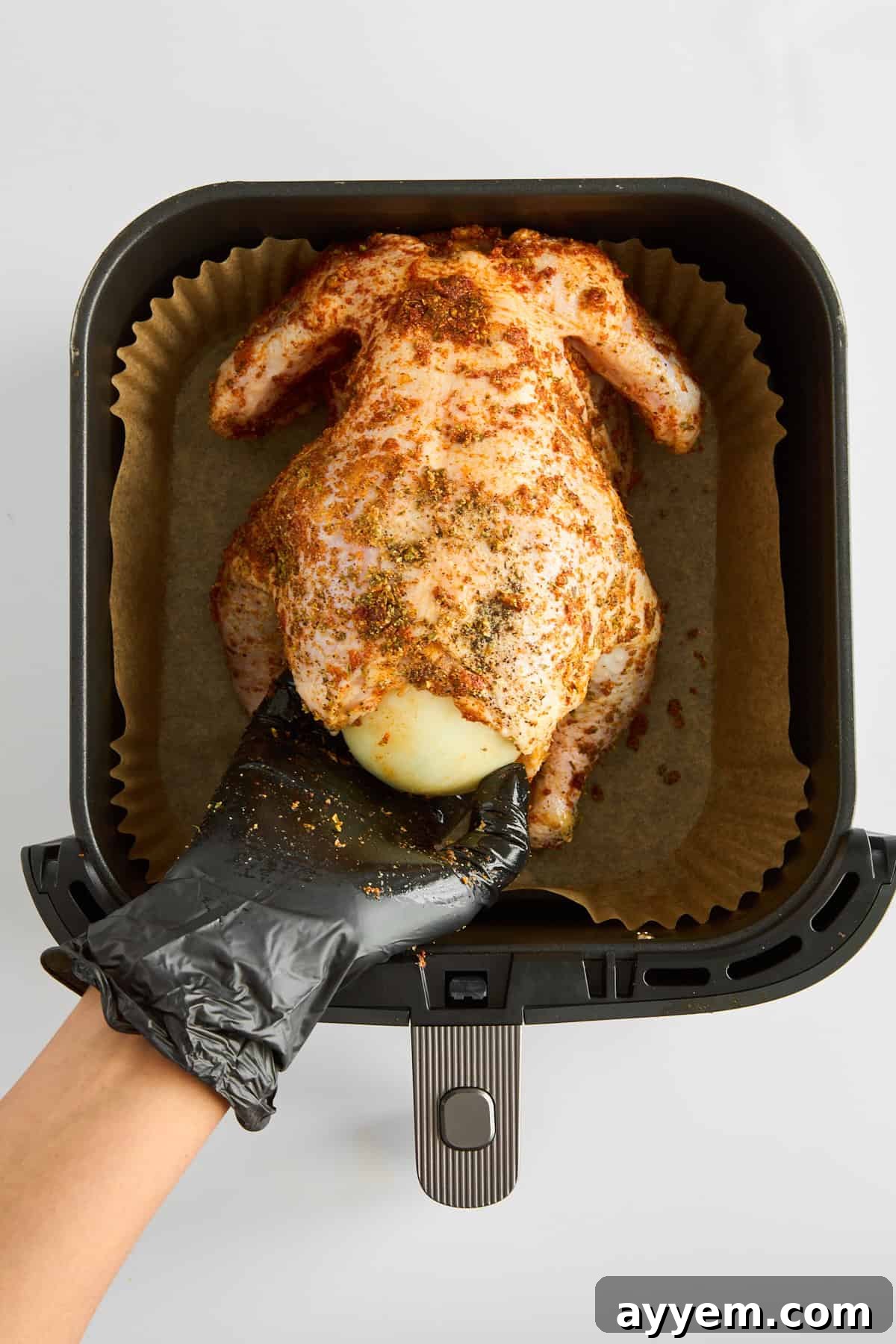 A gloved hand is stuffing a raw whole chicken with a halved onion, preparing it for the air fryer.