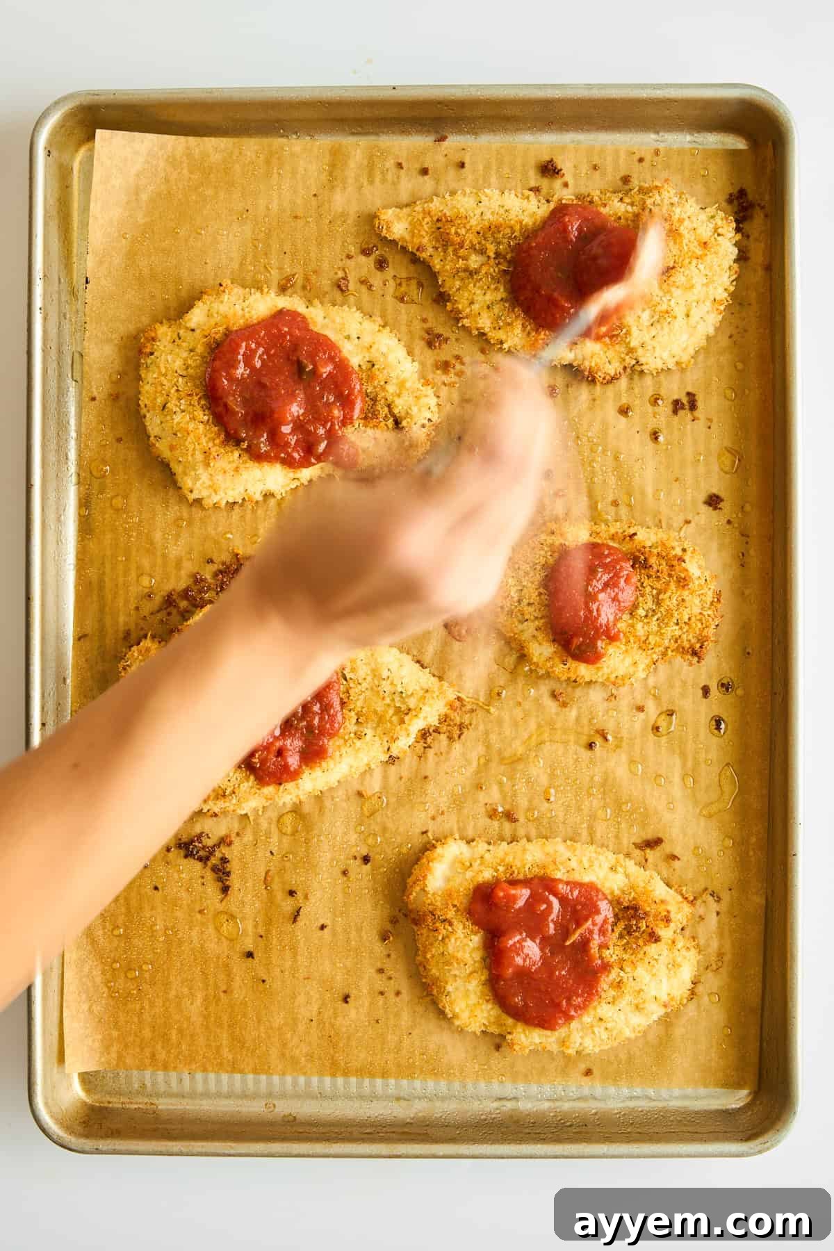 Marinara sauce being spooned evenly over partially baked breaded chicken breasts on a baking sheet.
