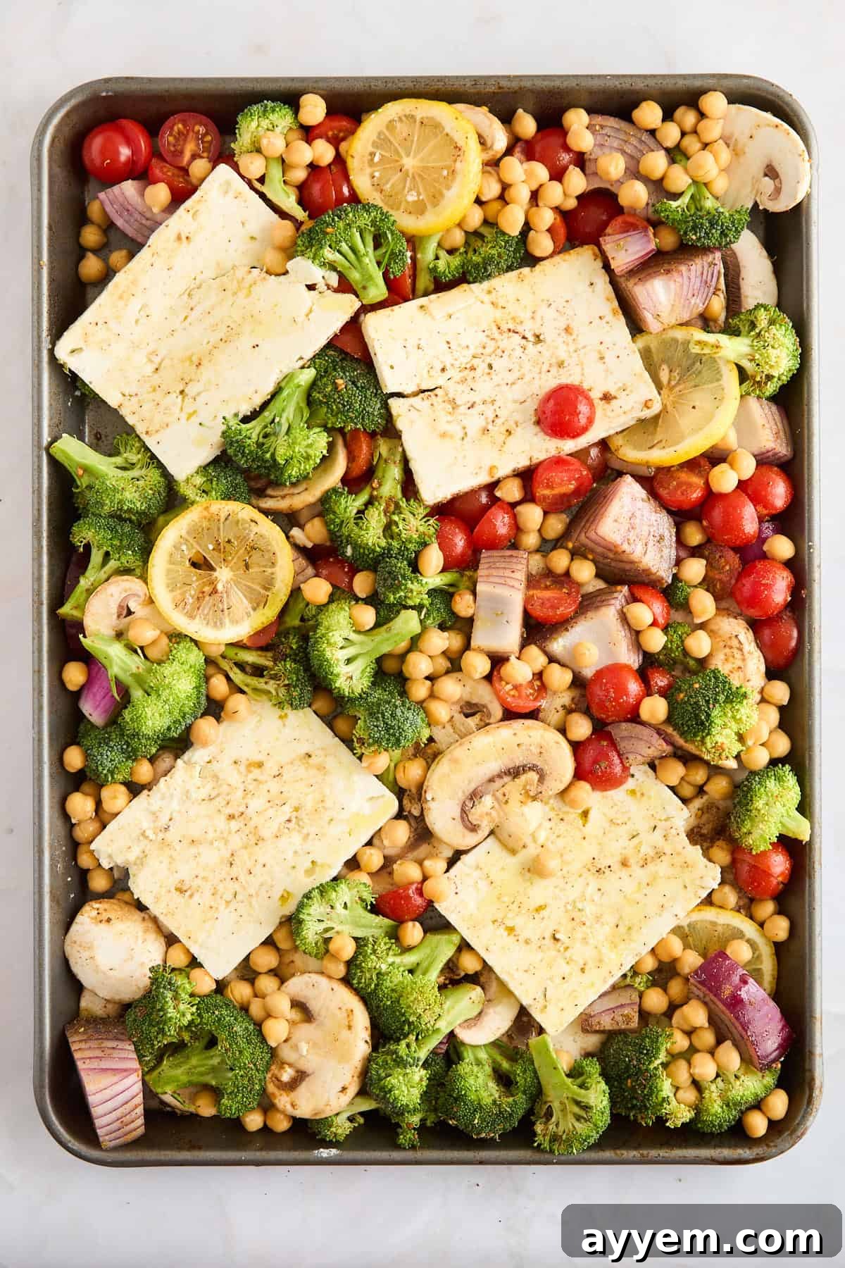 Seasoned raw vegetables, feta, and chickpeas uniformly spread on a baking sheet before roasting.