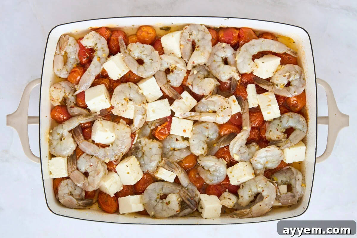 Raw shrimp and feta cubes nestled over baked tomatoes in a baking dish.