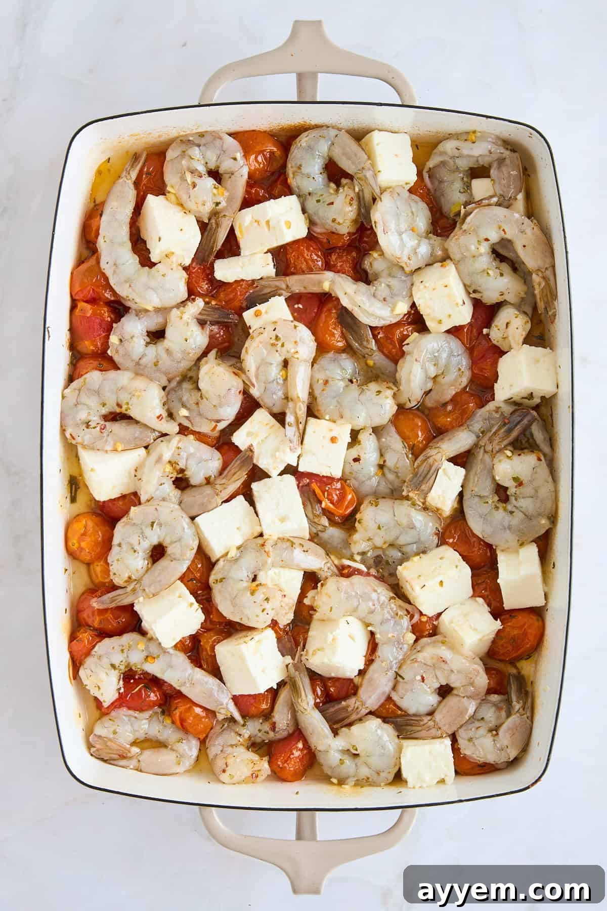 Baked tomatoes topped with feta cubes and raw seasoned shrimp, arranged in a baking dish.