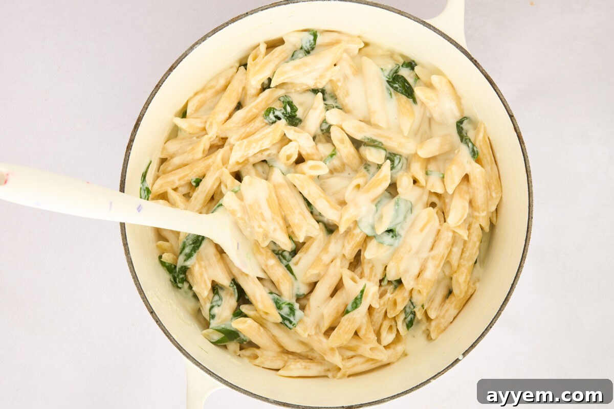 Penne pasta being tossed with cottage cheese sauce and spinach in a pot.