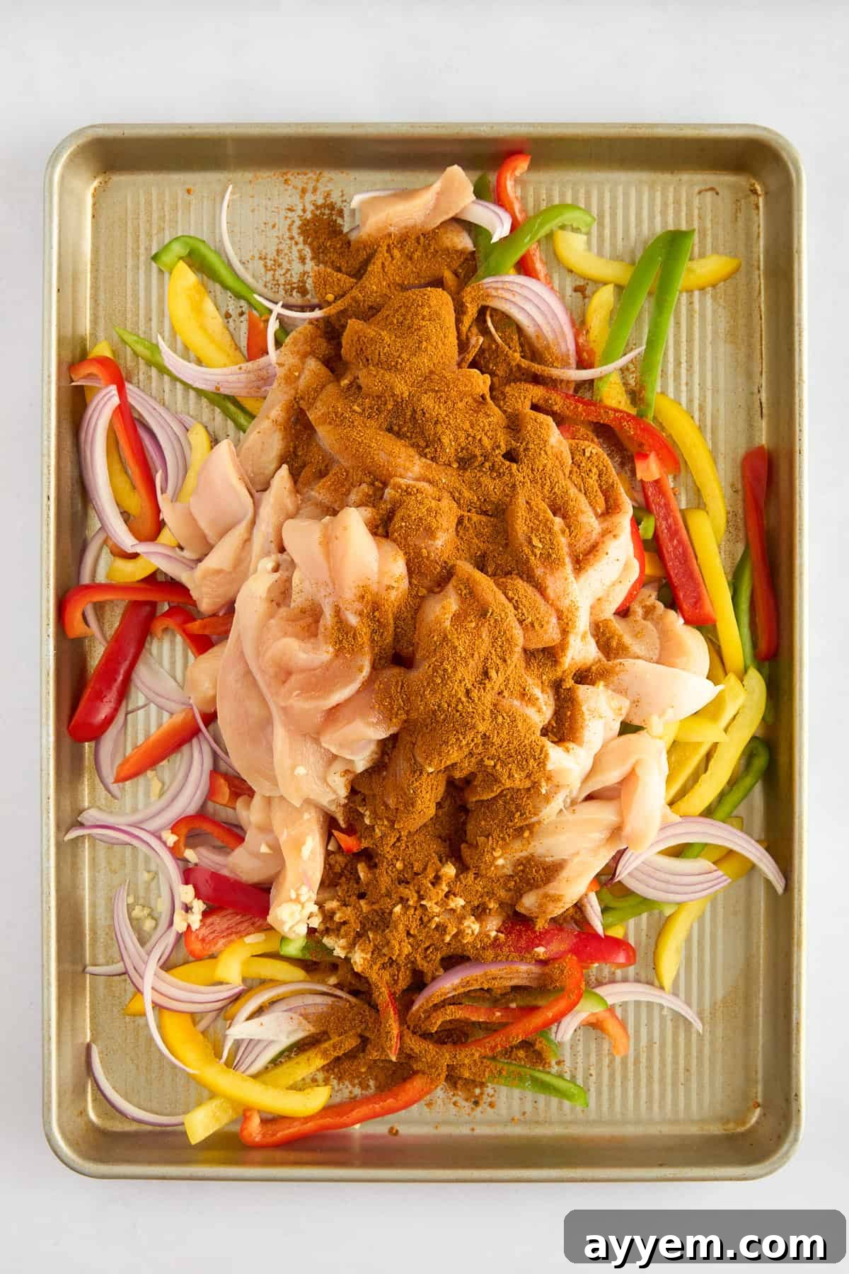 Raw chicken breasts, sliced red onions, and vibrant bell peppers arranged on a baking sheet, generously coated with aromatic fajita seasoning before baking.