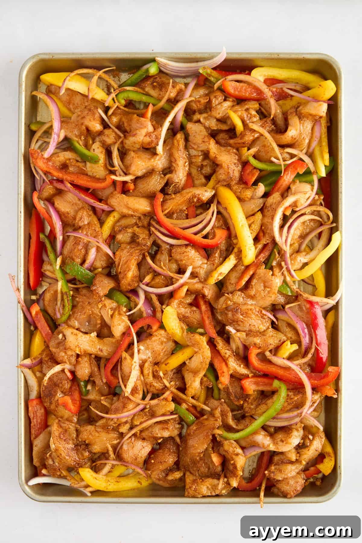 Unbaked fajita-seasoned chicken slices, colorful bell peppers, and red onions evenly spread on a baking sheet, ready for the oven.