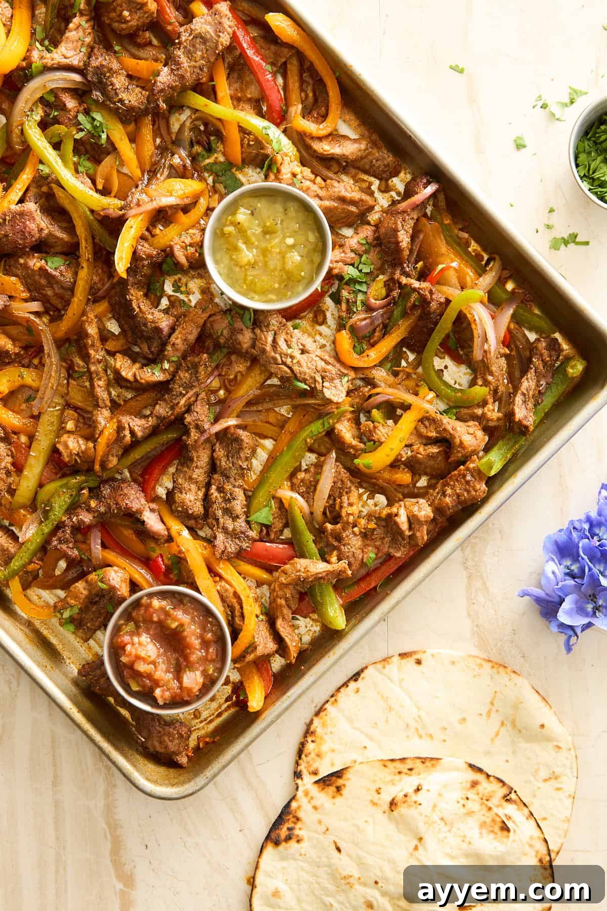 Effortless Sheet Pan Steak Fajitas 2 A baking sheet full of sizzling steak fajitas, with vibrant bell peppers and onions, served with sides of salsa verde and red salsa.