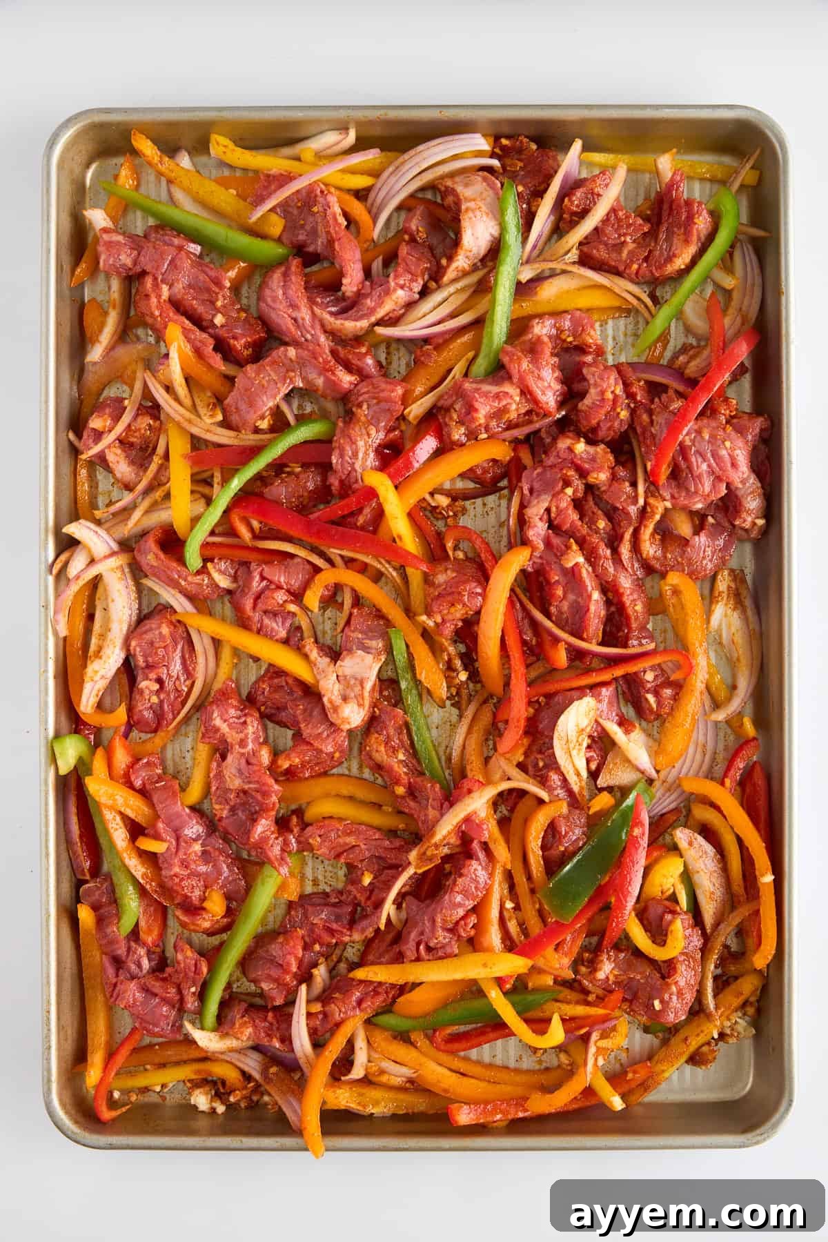 Effortless Sheet Pan Steak Fajitas 3 Raw fajita coated steak strips, bell peppers, and onions arranged on a baking sheet, ready for the oven.