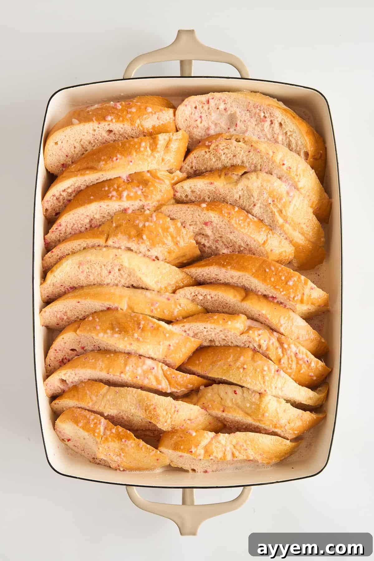 Thick slices of French bread absorbing a creamy, light pink strawberry custard in a baking dish, before baking.