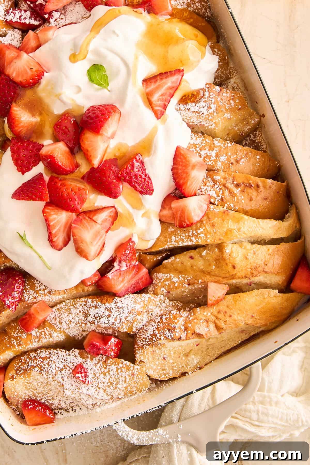 A baking dish of baked strawberry french toast, generously topped with fluffy whipped cream, fresh whole berries, and a drizzle of maple syrup.