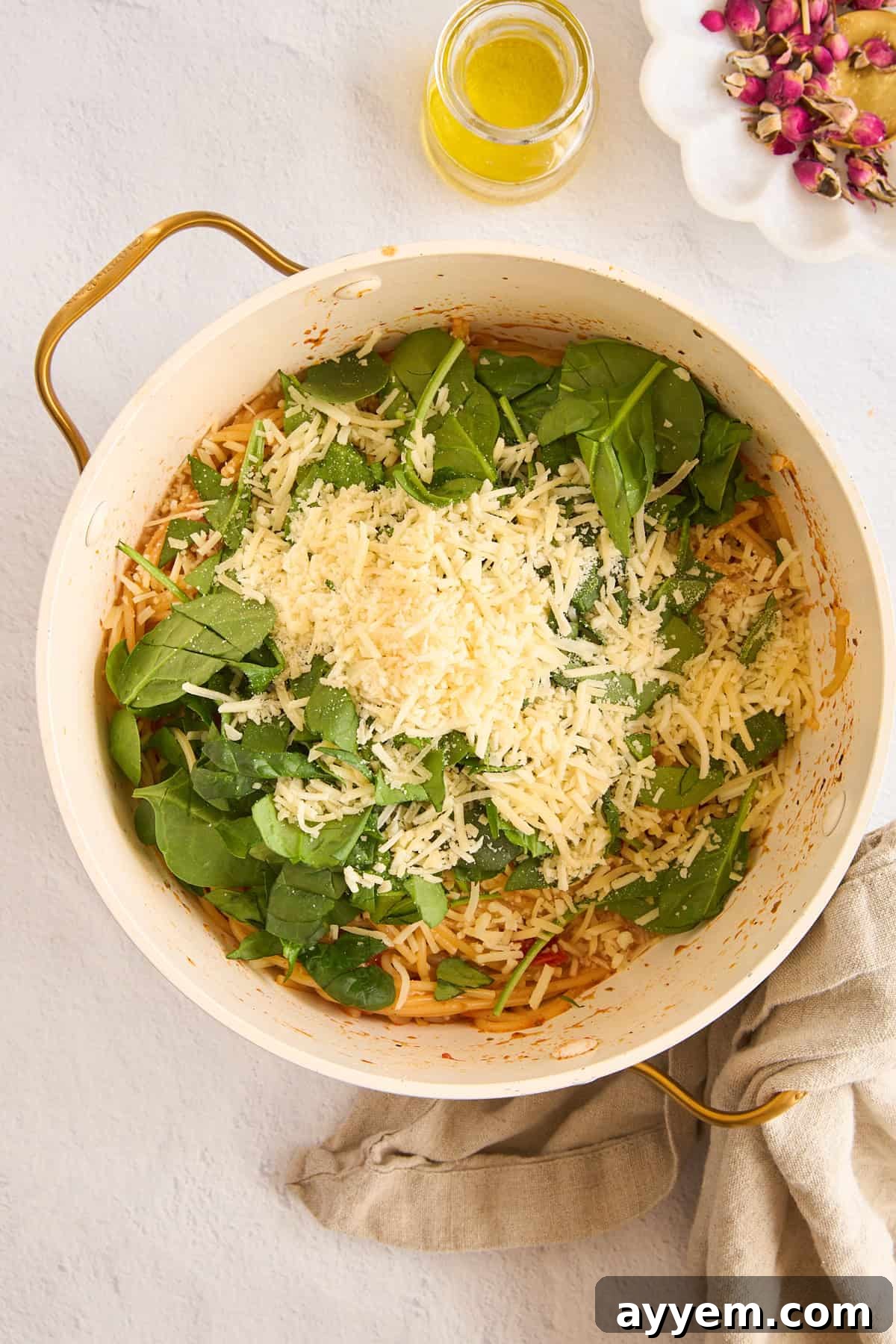 Fresh spinach and grated Parmesan cheese being added to a pot of cooked pasta with tomatoes and garlic, illustrating the final steps.