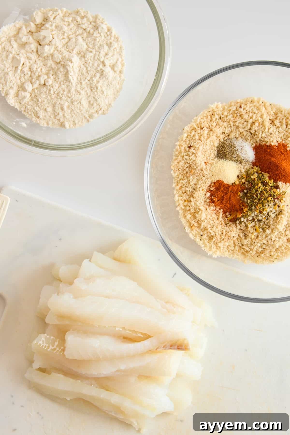 A bowl of flour, a bowl of seasonings, and raw cod fillets for homemade fish sticks.
