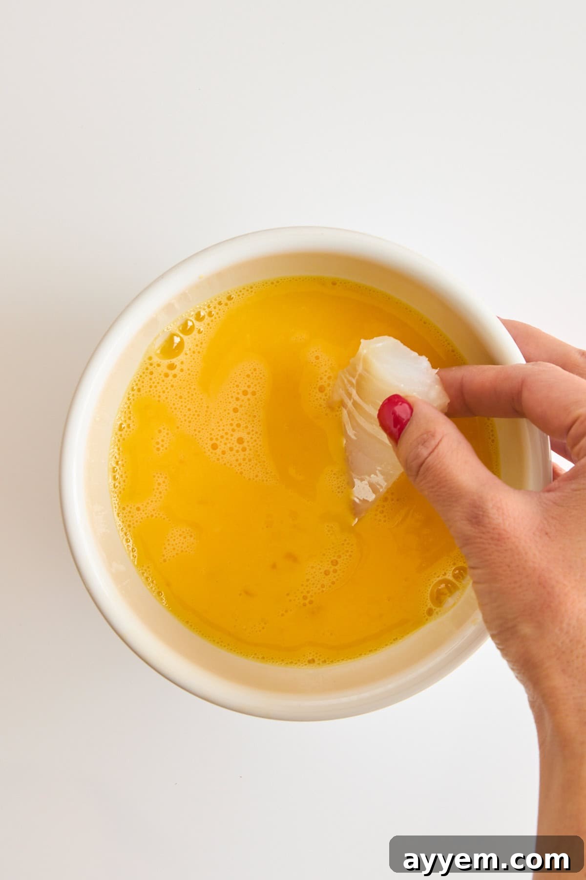 A raw cod piece being dipped into a bowl of whisked eggs for breading.