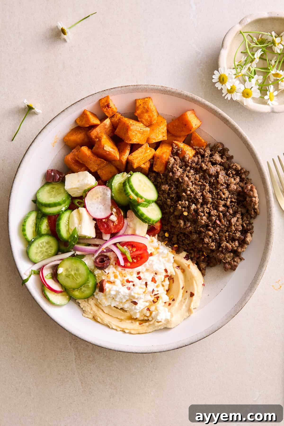 A close-up of a perfectly assembled Mediterranean ground beef bowl, showcasing the rich ground beef, golden roasted sweet potatoes, a colorful array of fresh vegetables, creamy feta, a dollop of hummus, and cottage cheese, all ready to be enjoyed.