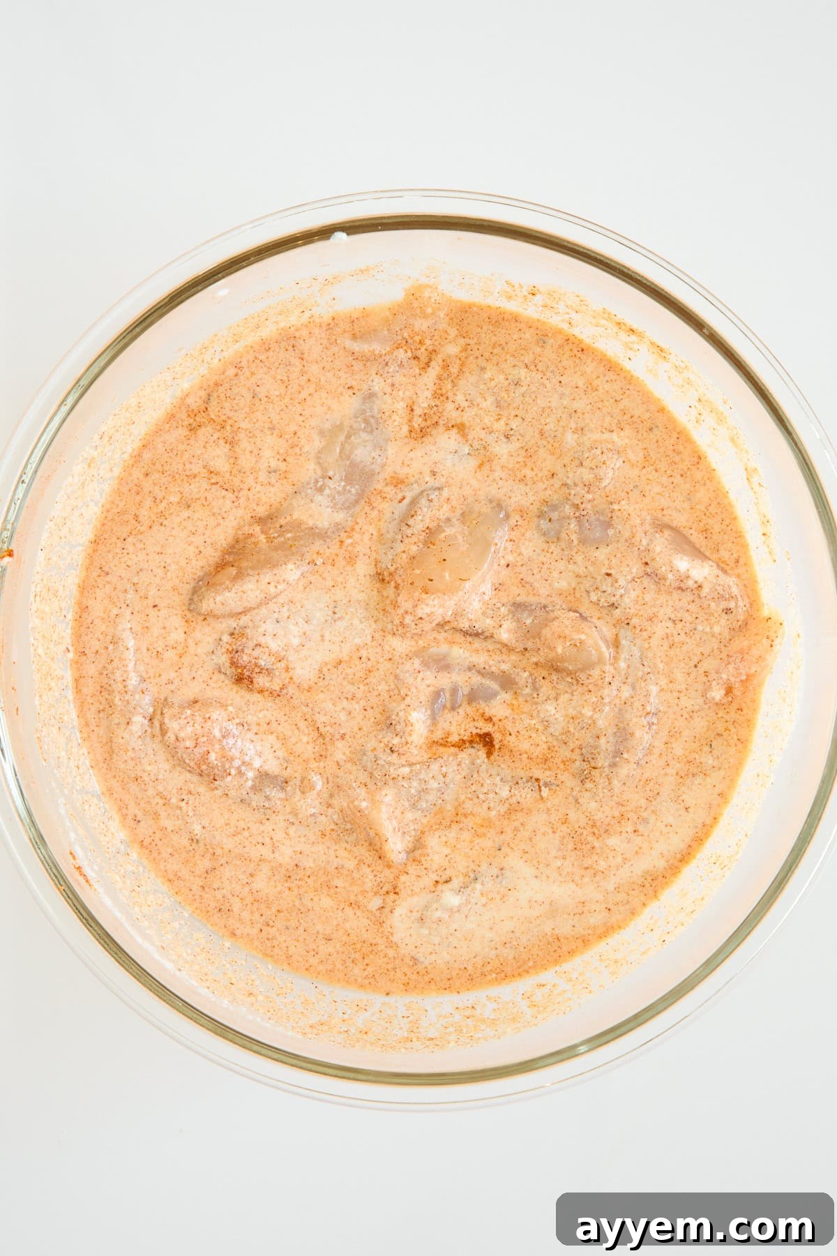 A clear glass bowl filled with pieces of chicken marinating in a creamy, orange-colored spice mixture, set against a plain white background.