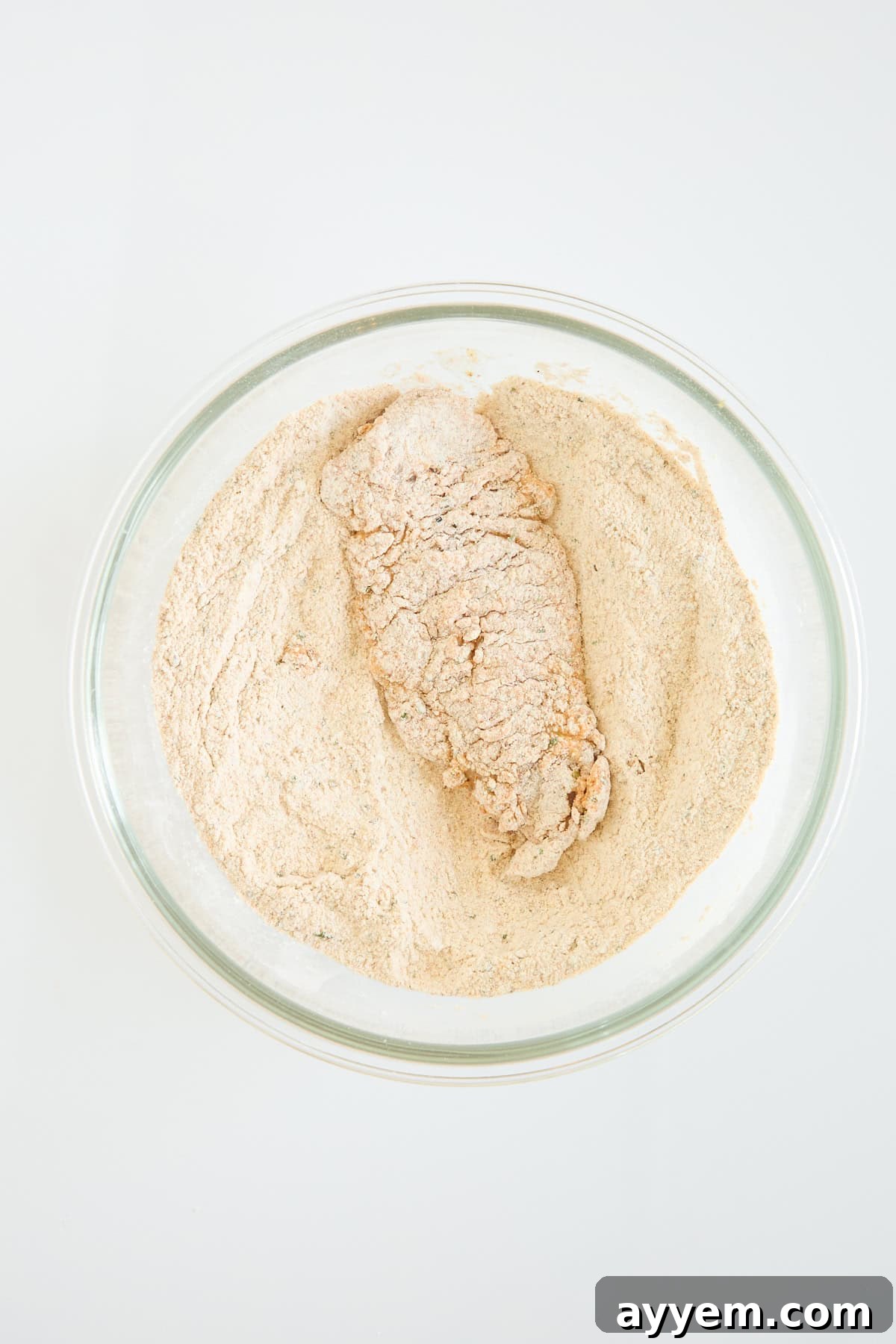 A piece of food, likely chicken, coated in flour and seasonings sits in a clear glass bowl filled with more flour mixture on a white surface.