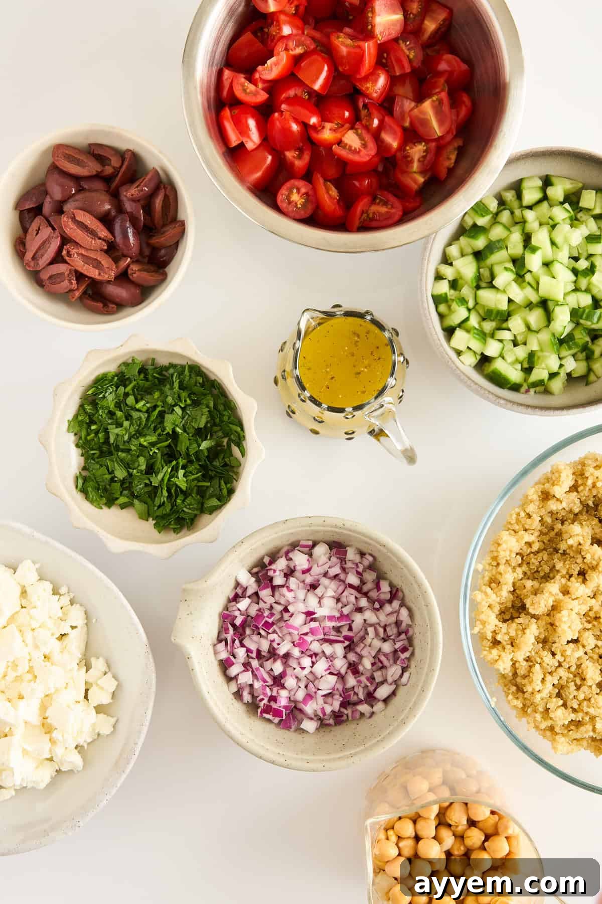 Bowls filled with neatly chopped tomatoes, cucumbers, red onions, fresh parsley, crumbled feta cheese, Kalamata olives, cooked quinoa, and chickpeas, with a small pitcher of yellow salad dressing arranged on a white surface, showcasing the fresh ingredients.