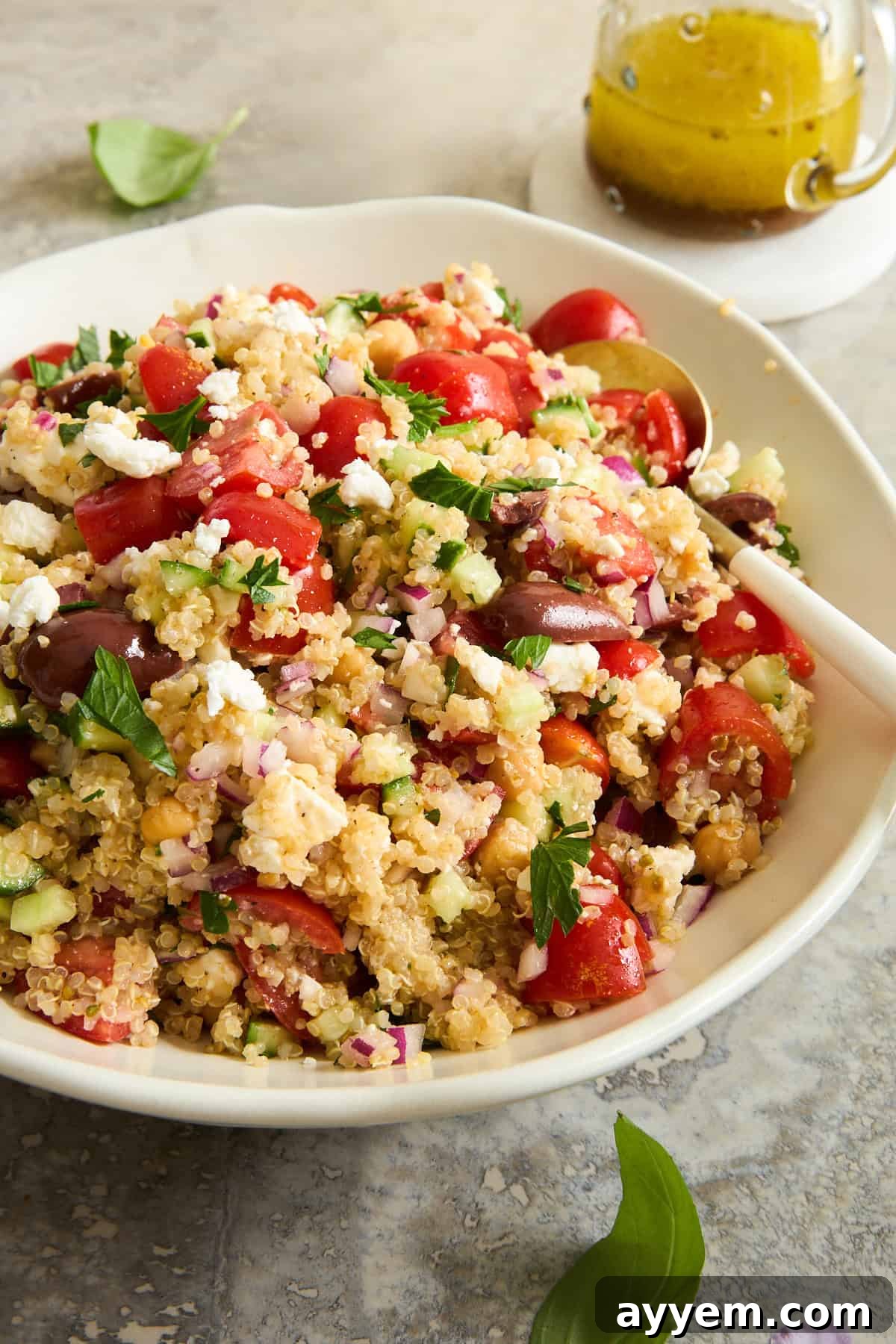 A close-up shot of a bowl of quinoa salad, beautifully composed with chopped tomatoes, cucumbers, red onions, Kalamata olives, fresh parsley, and crumbled feta cheese. A serving spoon rests in the bowl, with a small jar of vibrant dressing blurred in the background, suggesting its readiness to be served.