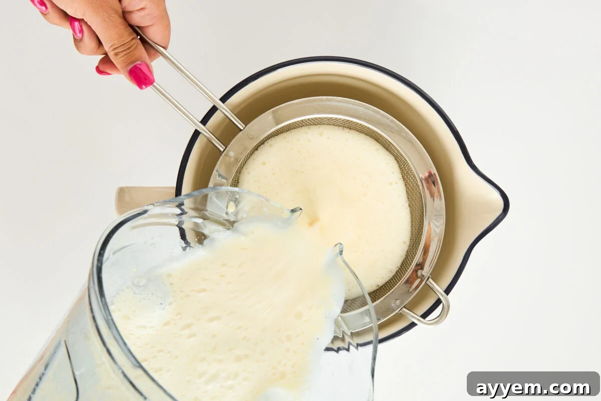 A hand holds a metal strainer over a cream-colored bowl while pouring a frothy liquid from a blender through the strainer. The person has pink nail polish. The background is white.