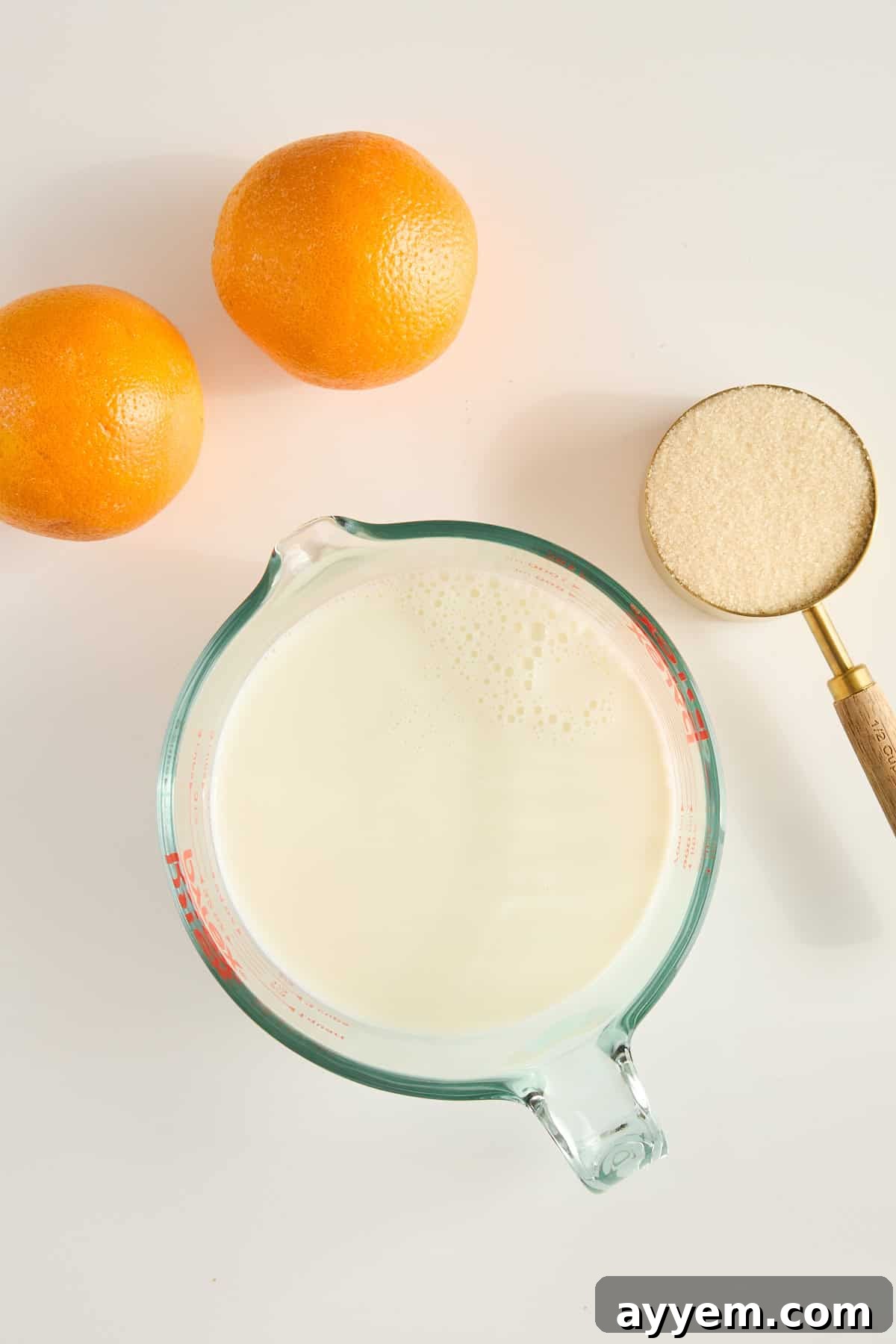 Two oranges, a glass measuring cup filled with milk, and a metal measuring cup with granulated sugar are arranged on a white surface, essential ingredients for an orange creamsicle drink.