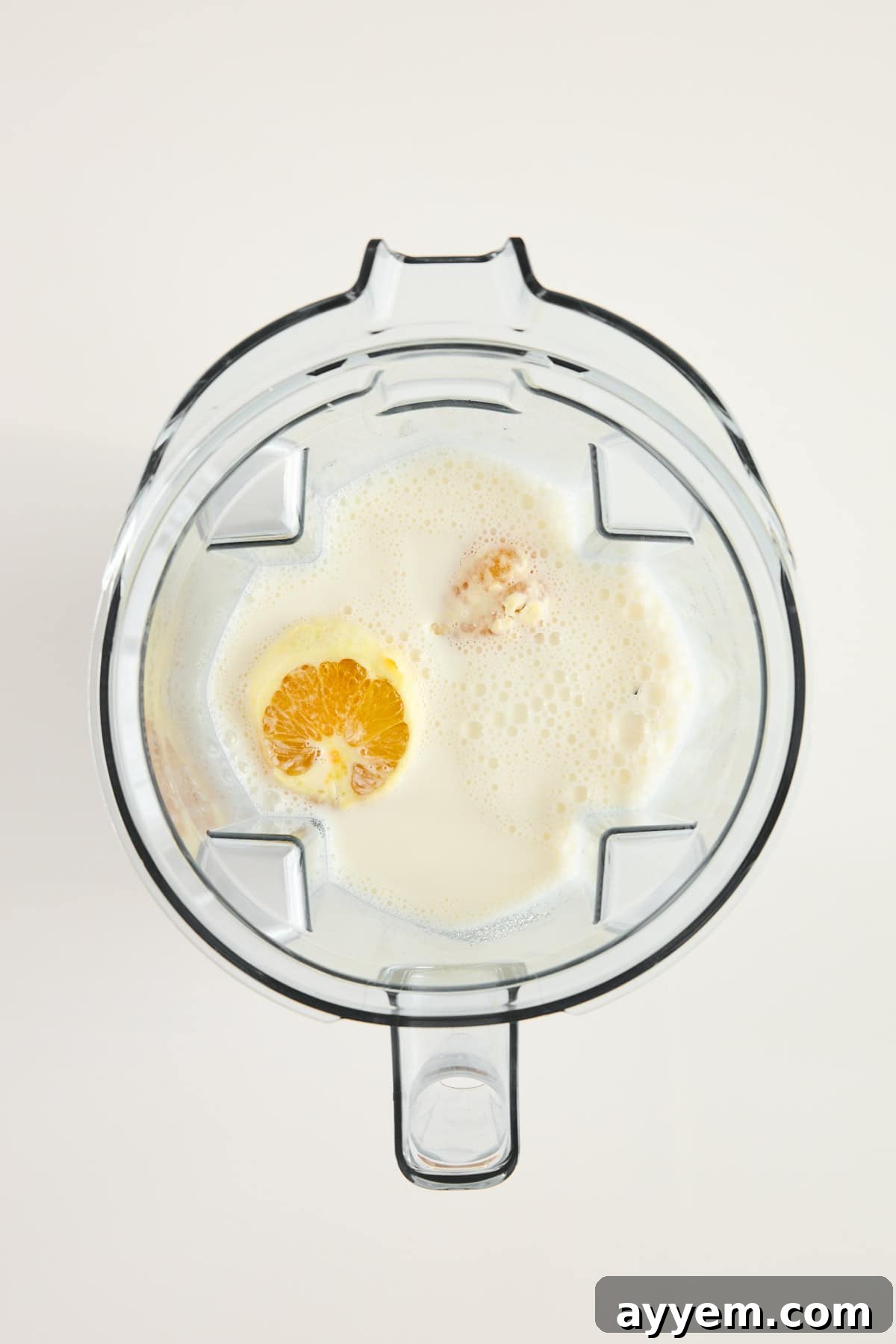 Top-down view of a blender containing milk, an orange half, and other ingredients, ready to be blended, on a white background, illustrating the first step of making an orange creamsicle.