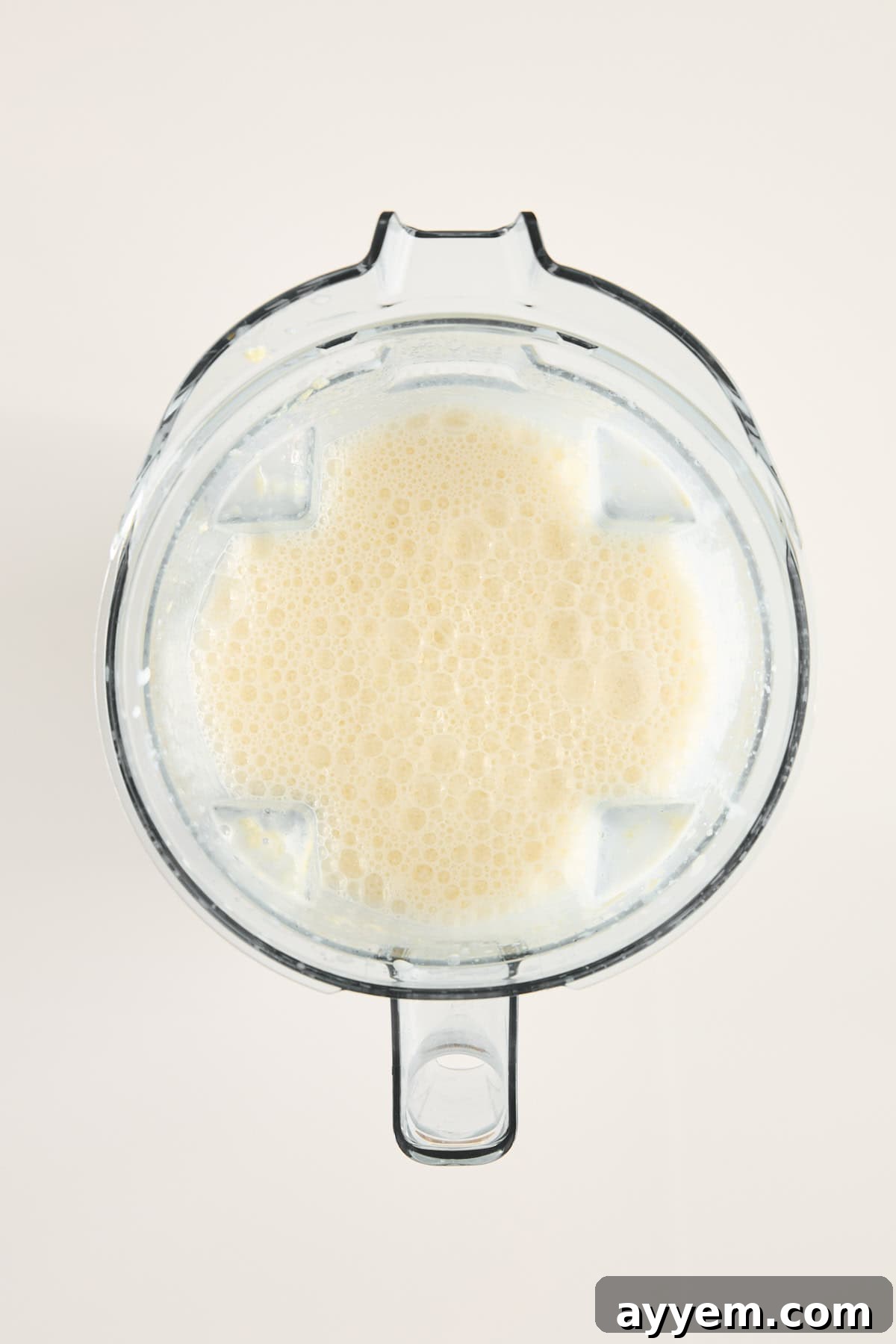 A top-down view of a blender filled with a light beige, frothy liquid, likely a smoothie or blended beverage, against a plain light background, showing the mixture after blending.