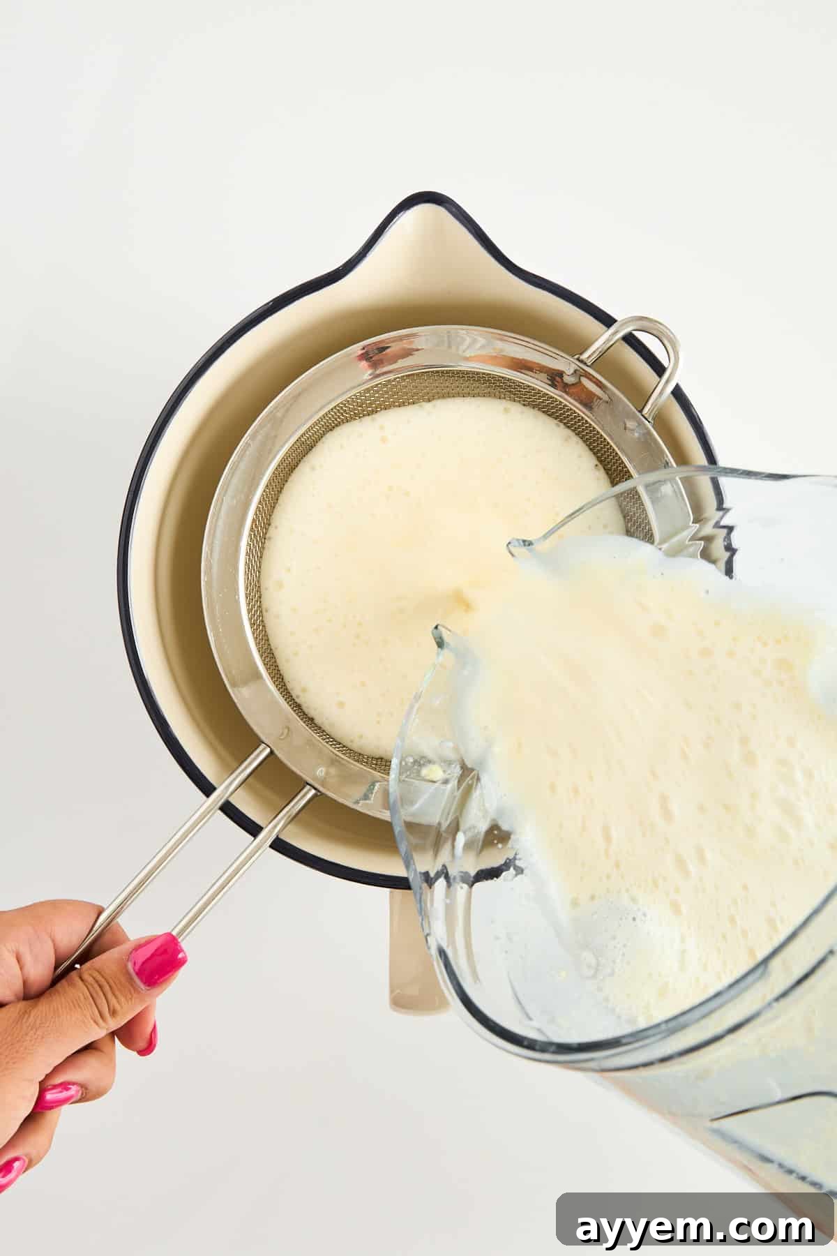 A hand holds a metal strainer over a cream-colored bowl while pouring a frothy liquid from a blender through the strainer. The person has pink nail polish. The background is white, demonstrating the straining process.