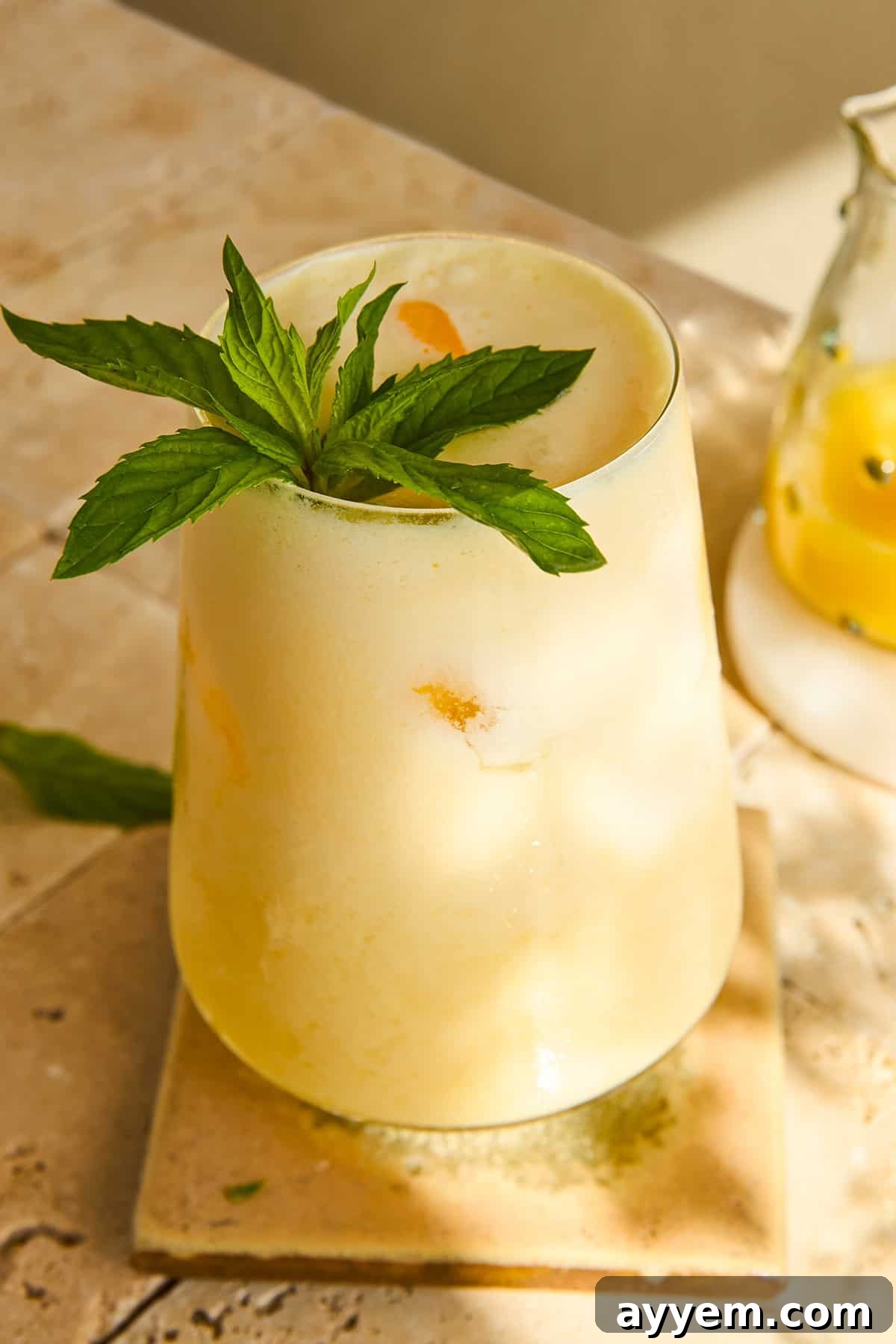 A frosty yellow drink garnished with a sprig of fresh mint sits on a square tile coaster, with sunlight casting soft shadows on the stone surface. A glass pitcher with yellow liquid is partly visible in the background, showcasing the finished orange creamsicle drink.