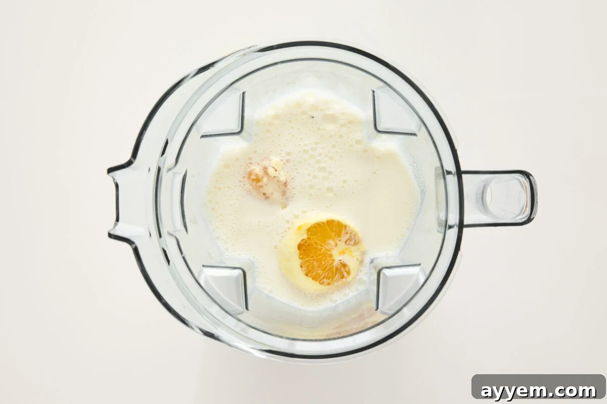 Top-down view of a blender containing milk, an orange half, and other ingredients, ready to be blended, on a white background.